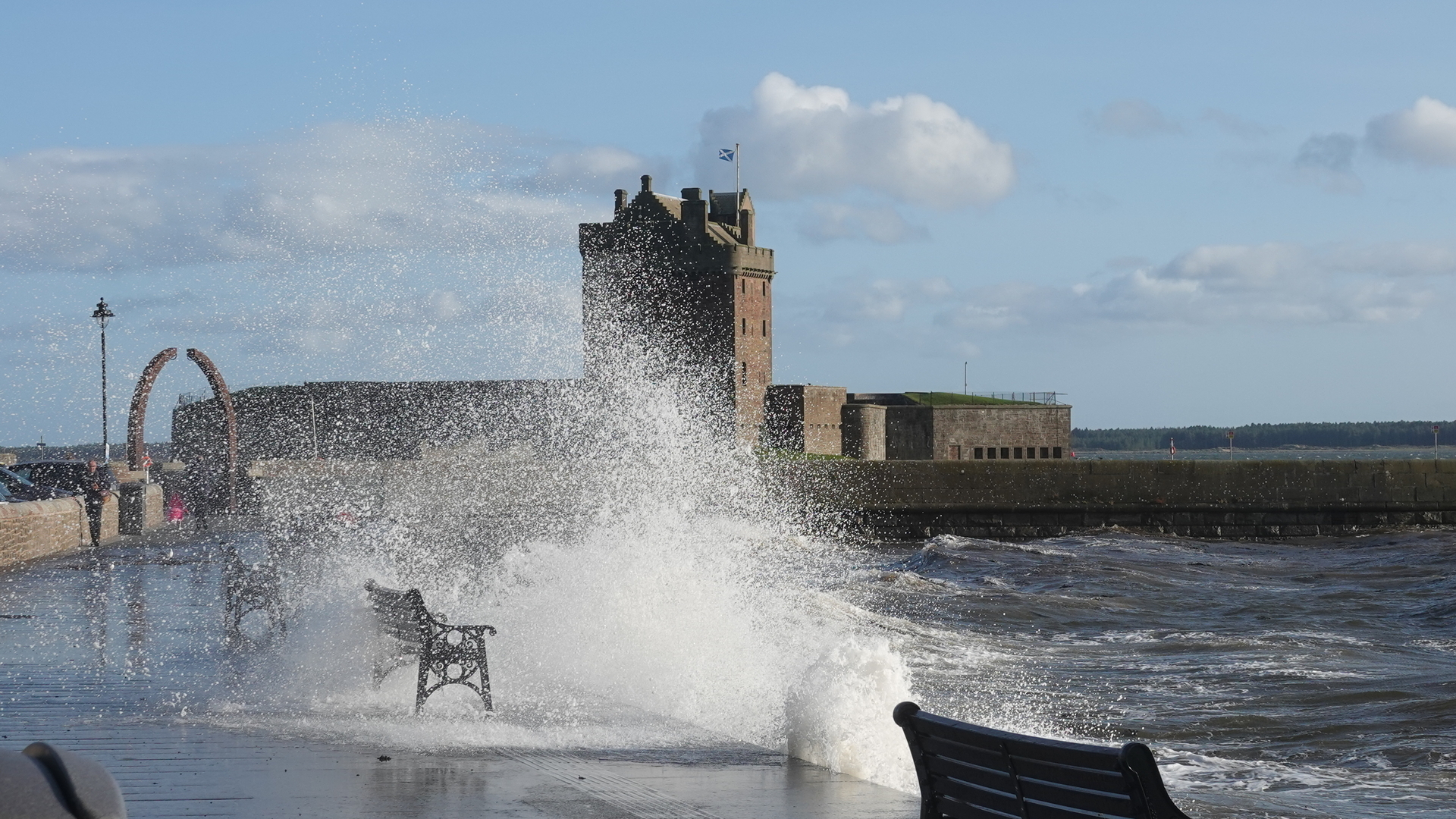 A seafront with the land to the left and benches running alongside it, and a very wild sea on the right throwing spray up onto the pavement and benches. In the background is an ornate arch sculpture, and beyond that an old castle with a tower in the middle. Behind is a slightly cloudy blue sky.