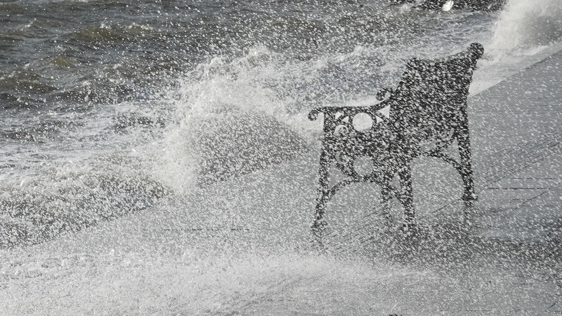 A dark bench sitting on a walkway beside the sea, totally drenched with seaspray which fills the image. The sea beyond it looks extremely wild.