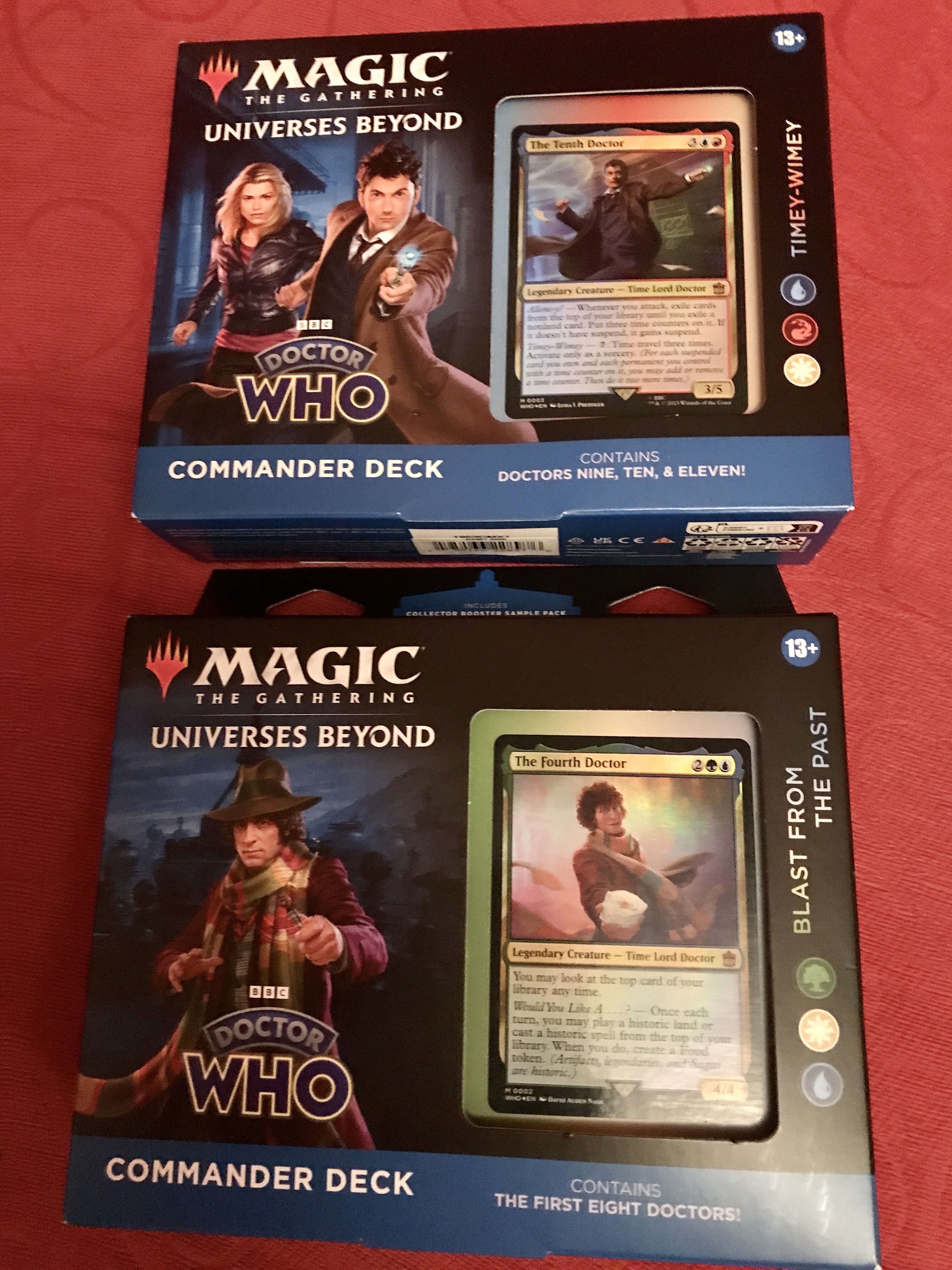 Two commander decks for Magic the Gathering, each in a blue cardboard box resting on a red sofa. The top box Timey Wimey features doctors 9-11 and shows Rose and the Tenth Doctor on the box. The deck below Blast From The Past features doctors 1-8 and shows Tom Baker’s Fourth Doctor in typical scarf, coat and hat costume.