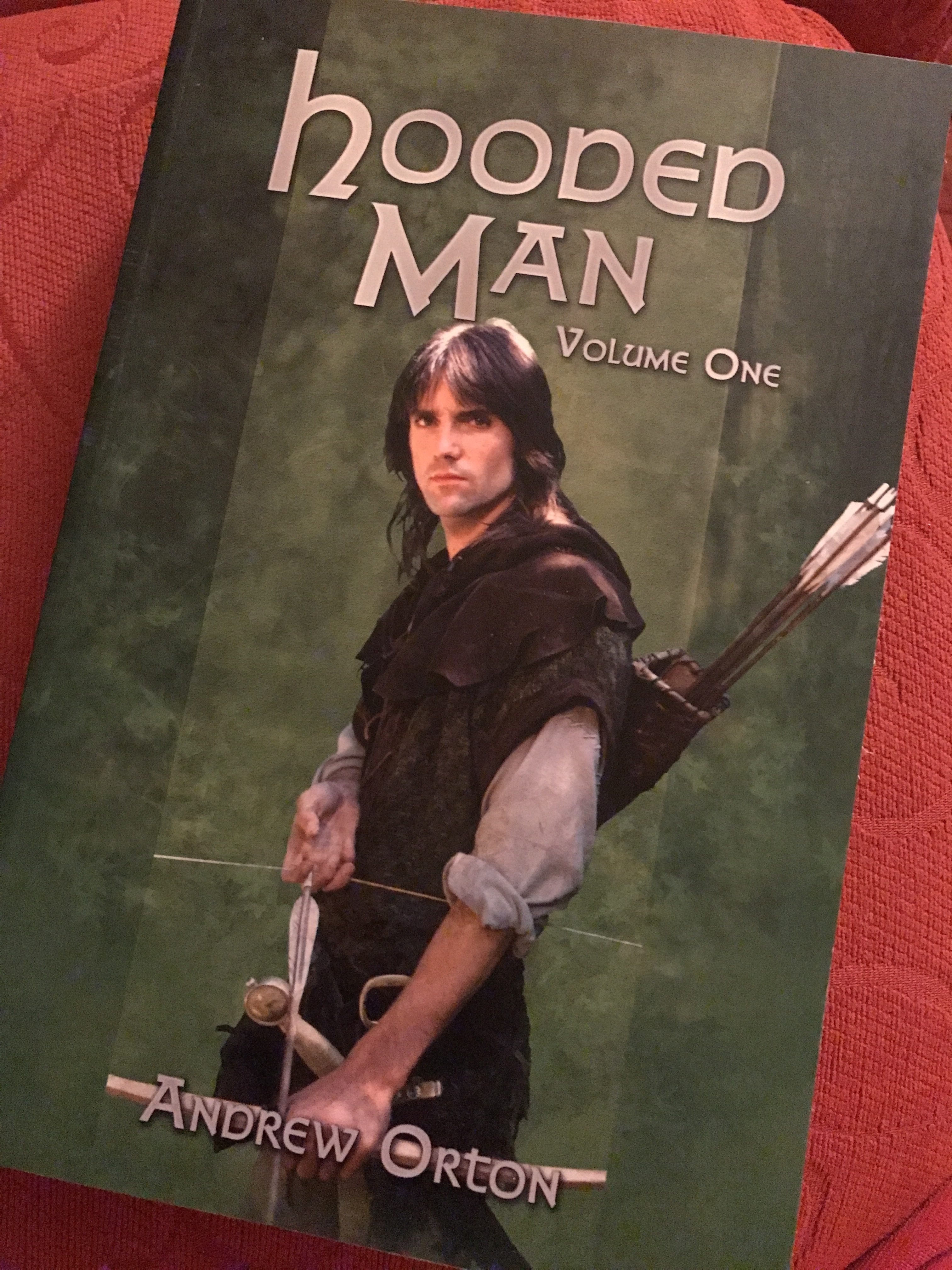 The book “Hooded Man volume one” resting on a red sofa. The book has a green cover, and shows Michael Praed’s Robin with bow and arrow.