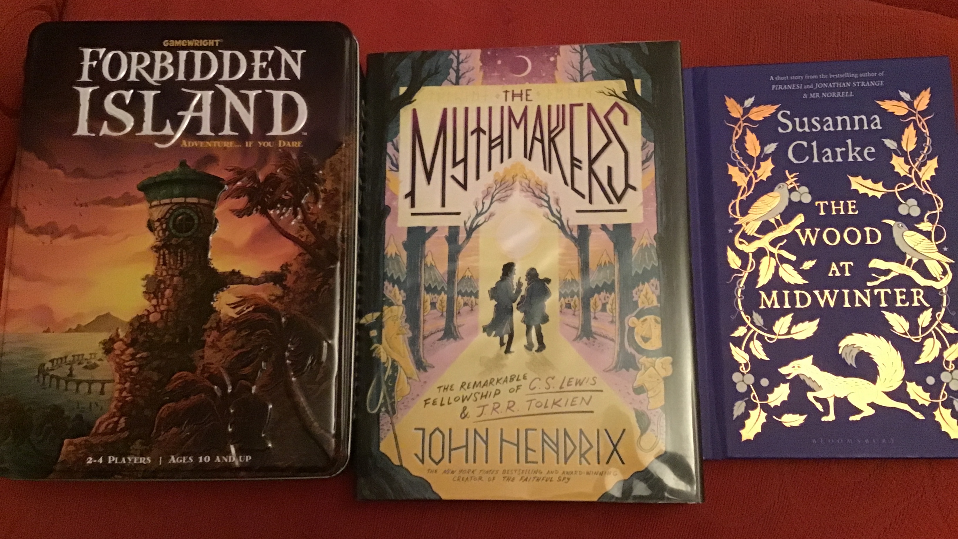 Three things resting beside each other on a red sofa. From left to right: 1/ the Forbidden Island board game in a brightly coloured tin container; 2/ The Mythmakers by John Hendrix graphic novel, featuring Tolkien and Lewis on the cover; and 3/ The Wood at Midwinter by Susanna Clarke, a small hardback book with a gold design of birds and woodland animals and leaves against a dark blue background.
