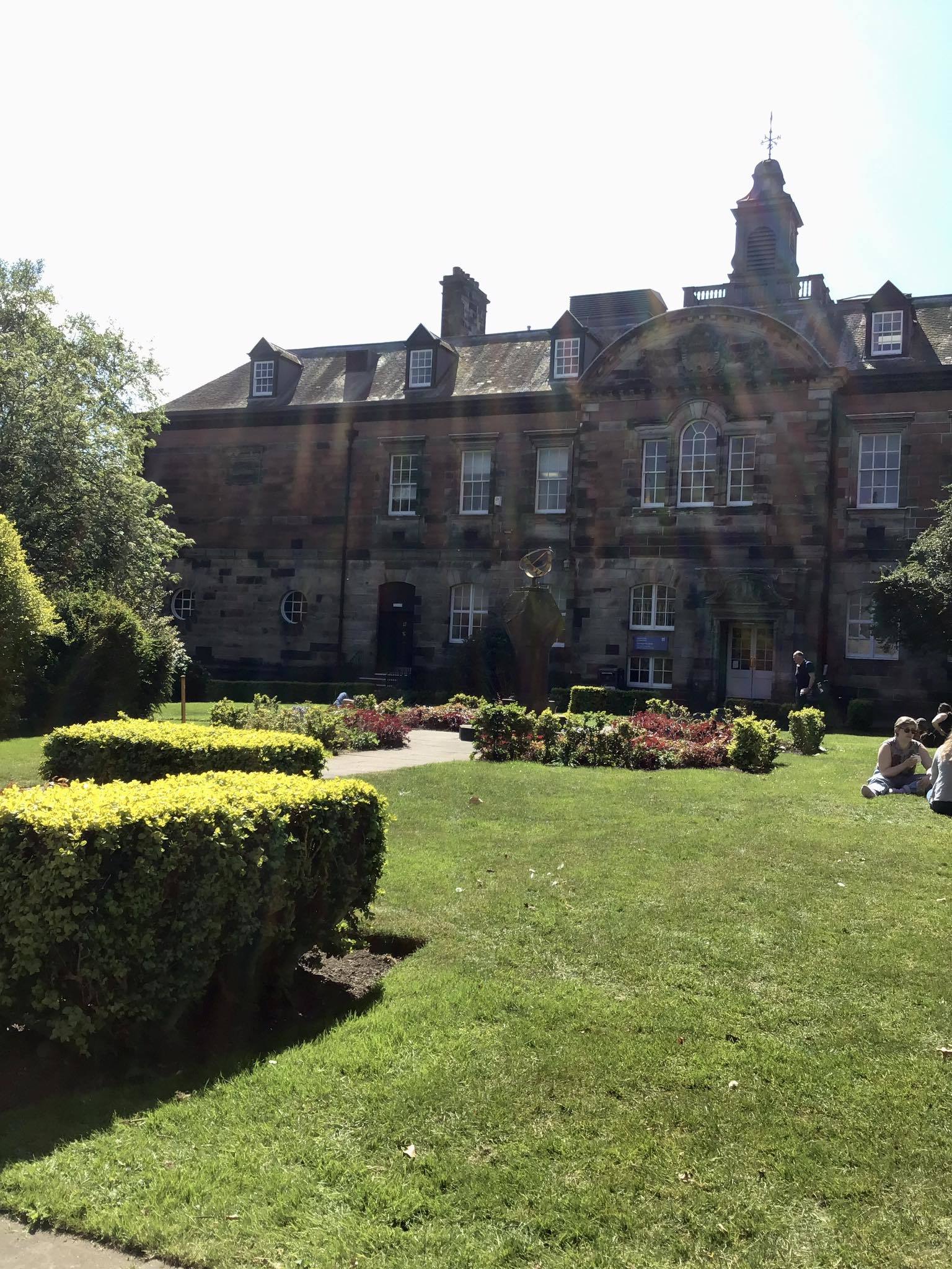 Photo of a sunny Geddes Quadrangle at Dundee University Grass and paths in between in front of an old red brick ornate building. Small hedges and beds of flowers are spread around. An ornate sculpture is just visible in the middle distance before the building. Sun was beaming down and the picture includes diagonal sun rays, especially visible across the building.